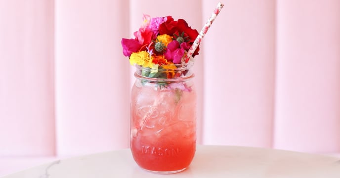 A pink drink inside of a mason jar that has a ton of pink flowers on the top of it, with a pink straw coming out. On of a counter with a pink background. 