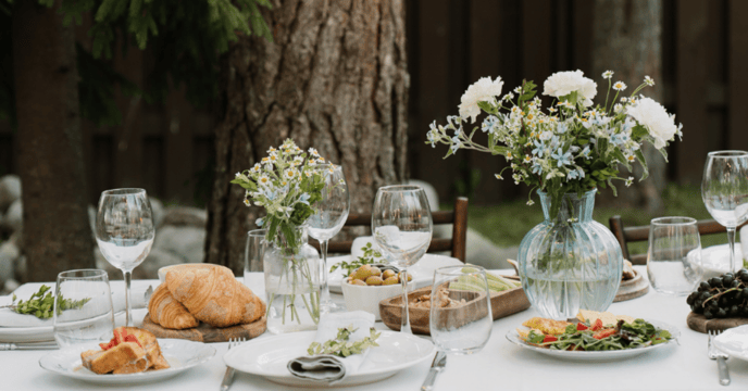 An outdoor dining set up. There are flowers in the middle of the table with organic food and white plates and white tablecloth. 