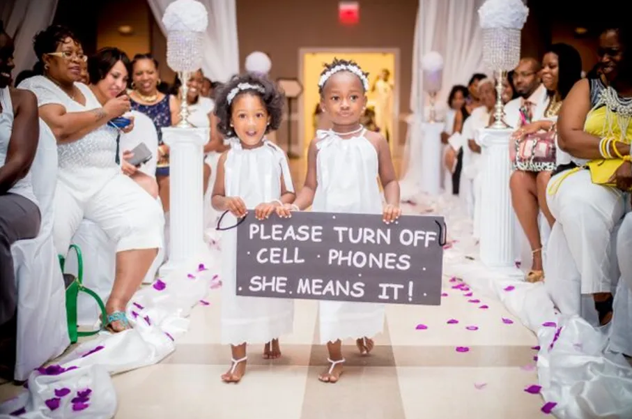 Two girls walking down the aisle holding a sign that says, "Please turn off cell phones she means it!"